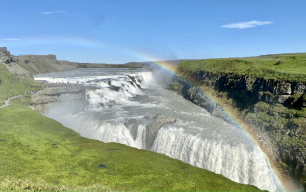 Picture of view at Gullfoss Waterfall in sunny day. Small rainbow visible over the waterfall and group of tourist in a far distance.